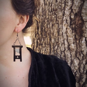 May include: A pair of silver earrings with a black guillotine charm. The guillotine is made of metal and has a silver blade. The earrings are hanging from a silver chain.