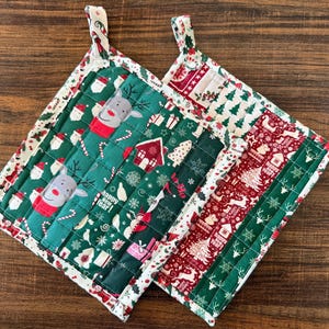 May include: Two square quilted pot holders featuring Christmas-themed fabric with reindeer, Santa Claus, gift boxes, and trees. The patterns include red, green, and white colors with text that reads Happy New Year and Ho Ho Ho.