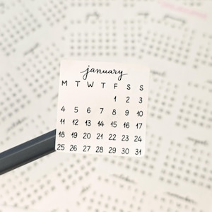 Könnte beinhalten: Ein kleiner, quadratischer Kalenderaufkleber für Januar mit handschriftlicher Schrift. Die Wochentage sind abgekürzt und die Daten sind übersichtlich angeordnet. Ein Bleistift ist teilweise im Vordergrund zu sehen, und andere Kalenderseiten sind im Hintergrund.