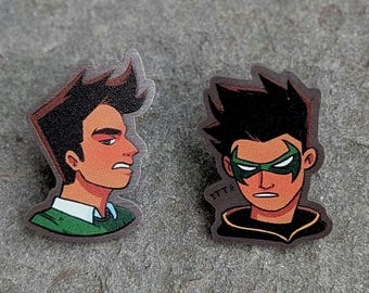 Può includere: Due spille in stile cartone animato con personaggi maschili. Una spilla mostra un profilo laterale con capelli castani, una camicia verde e un viso arancione. L'altra spilla raffigura un personaggio con una maschera verde, capelli neri e un abito nero e oro.