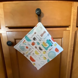 Könnte beinhalten: Ein weißes Küchentuch mit einem bunten Muster aus Küchenutensilien und -geräten. Das Tuch hängt an einem Haken an einer Holztür.