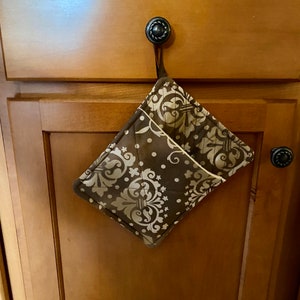 May include: A brown and cream patterned pot holder with a loop for hanging. The pot holder is hanging on a cabinet door.