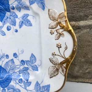 May include: A white porcelain serving dish with a blue and gold floral design. The dish has a scalloped edge and a raised rim. The floral design features blue leaves and berries on a white background. The rim of the dish is accented with gold.