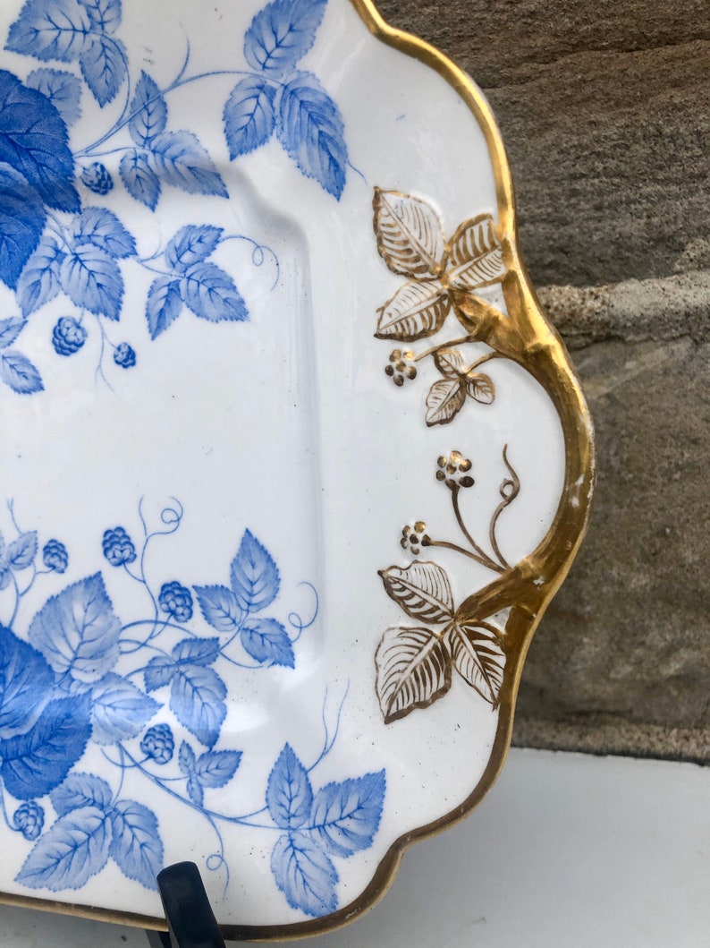 May include: A white porcelain serving dish with a blue and gold floral design. The dish has a scalloped edge and a raised rim. The floral design features blue leaves and berries on a white background. The rim of the dish is accented with gold.