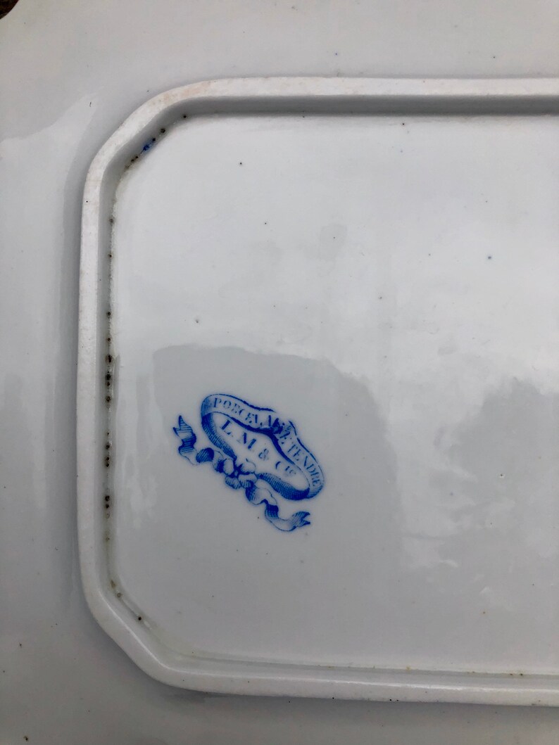 May include: A close-up of a blue and white porcelain mark on a white plate. The mark reads "Porcelain de Tendee L.M. & Cie."