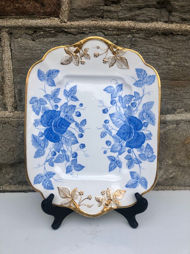 May include: A white rectangular serving platter with a blue and gold floral design. The platter has a scalloped edge and is decorated with a repeating pattern of blue leaves and berries with gold accents.