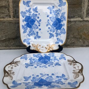 May include: Two white porcelain serving platters with blue and gold floral designs. The platters have a square shape with rounded corners and a raised rim.