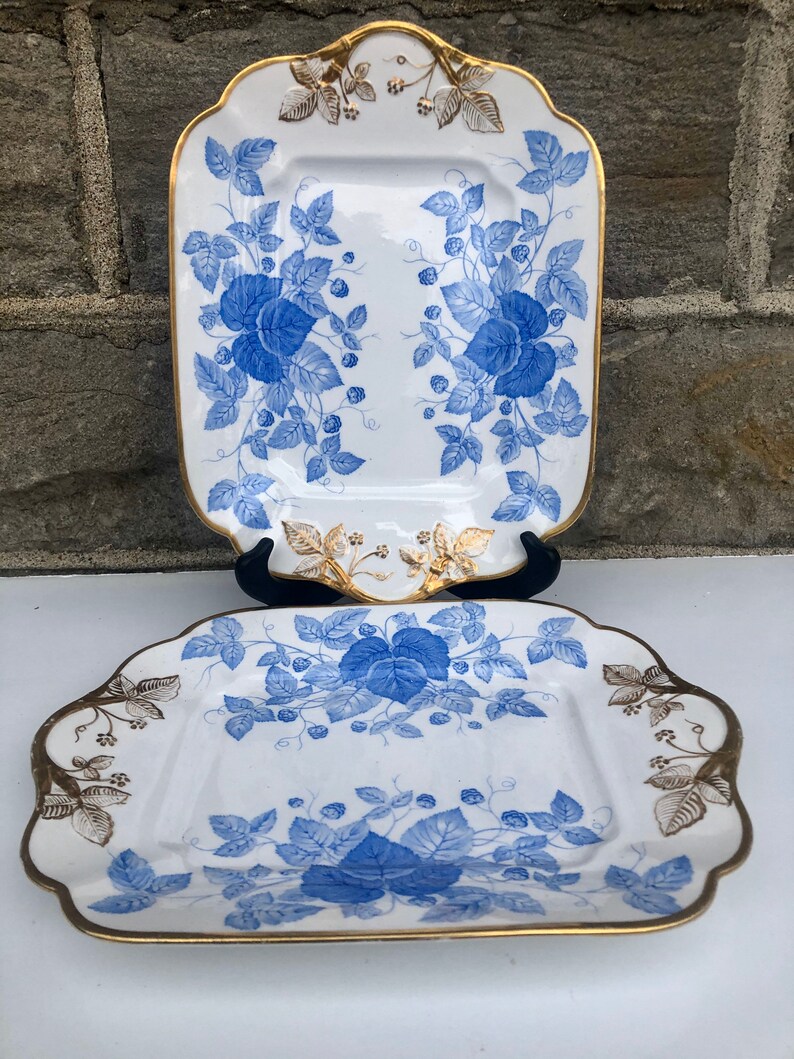 May include: Two white porcelain serving platters with blue and gold floral designs. The platters have a square shape with rounded corners and a raised rim.