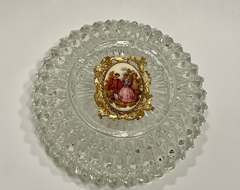 May include: A clear glass trinket dish with a raised, textured design. The dish features a gold-toned oval frame with a porcelain cameo of a couple in a garden setting.