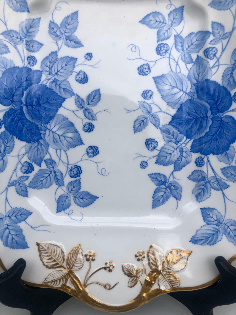 May include: A white porcelain plate with a blue and gold floral design. The plate has a scalloped edge and is decorated with a repeating pattern of leaves and berries.