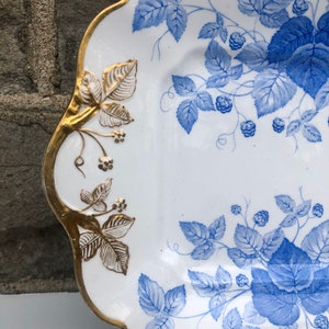 May include: A white porcelain serving platter with a blue and gold floral design. The platter has a scalloped edge and is decorated with a repeating pattern of blue leaves and berries.