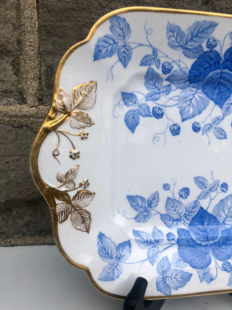 May include: A white porcelain serving platter with a blue and gold floral design. The platter has a scalloped edge and is decorated with a repeating pattern of blue leaves and berries.