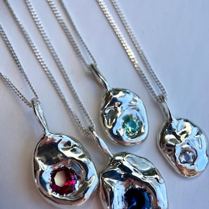 May include: Four silver pendant necklaces. Each necklace has an irregularly shaped pendant with a different coloured gemstone: red, blue, green, and clear. The pendants hang from delicate silver chains.