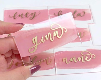 May include: Clear acrylic place cards with gold calligraphy names over a pink painted brushstroke. The name "Gina" is in focus, with other names like "Ency" and "Anne" visible. Ideal for weddings or events.