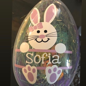 Peut inclure: Oeuf de Pâques en plastique transparent avec un motif de visage de lapin blanc et rose. L'œuf porte le nom "Sofia" en écriture blanche et est rempli d'herbe décorative verte et violette. Le texte "Comes in green blue pink purple" est en haut.