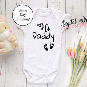 Op de afbeelding: Witte babybody met de tekst "Hi Daddy" in zwart schrift, vergezeld van twee zwarte voetafdrukken. Een pluchen konijn en een boeket tulpen staan ook in de afbeelding. De tekst "Digital Dreams Custom Printing" staat in de afbeelding.