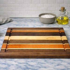 May include: A rectangular wooden chopping board with a striped design in shades of brown, orange, and tan. The board has a juice groove and is surrounded by kitchen items, including a bowl of salt, a bottle of oil, and a small plant.