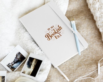 May include: A white notebook with a brown inscription that reads "In My Bookish Era". The notebook is lying on a white fuzzy blanket with a blue pen and a camera.