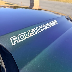 Puede incluir: Primer plano del capó de un coche con un acabado degradado azul y verde. El texto "ROUSHCHARGED" se muestra en letras blancas con contorno en el capó. El capó refleja el cielo y el entorno.