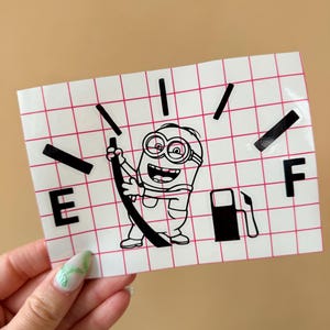 Puede incluir: Una pegatina blanca con un patrón de cuadrícula que presenta un personaje de dibujos animados Minion sosteniendo una manguera de bomba de gasolina. La pegatina imita un indicador de combustible, con "E" y "F" que indican vacío y lleno.