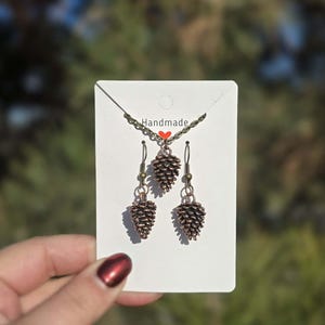 Puede incluir: Un conjunto de joyas hechas a mano con un collar y pendientes. El conjunto incluye un collar con colgante y dos pendientes a juego, todos con un diseño de piña. Las joyas se exhiben en una tarjeta blanca con la palabra "Handmade" y un pequeño corazón rojo.