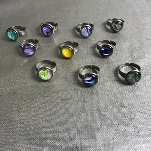 May include: A collection of ten silver rings with colorful, abstract designs on the top. The rings are arranged in two rows on a gray textured surface.