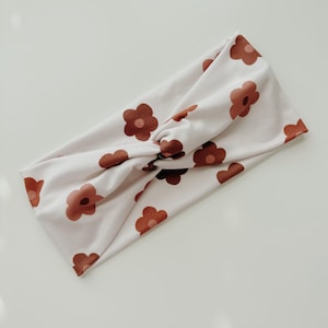 May include: A white headband with a brown floral pattern. The headband is made of a soft, stretchy fabric and is designed to be worn as a knotted headband.