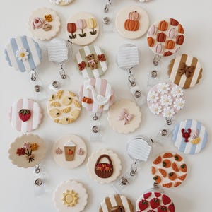 May include: A collection of circular badge reels featuring various decorative designs including striped patterns, flowers, pumpkins, fruit, animals, and seasonal motifs. Each badge reel has a retractable cord and a clear plastic clip for attachment.