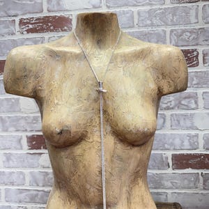 May include: A silver-toned necklace with a pendant, displayed on a mannequin. The pendant appears to be a stylized bird or wing shape. The necklace hangs down the mannequin's torso, showcasing its length and design. The background is a brick wall.