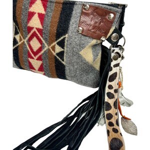 May include: A gray and brown patterned clutch with a black fringe and a brown leather patch with silver studs. The clutch has a zipper closure and a long, black and white spotted strap with a silver charm.