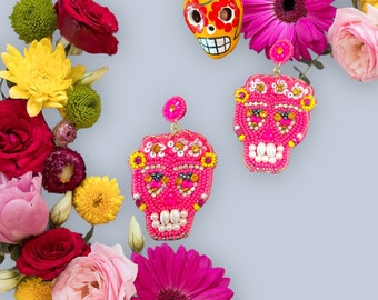 May include: A pair of pink beaded skull earrings with gold accents. The earrings are surrounded by colorful flowers.