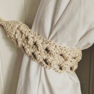 May include: A close-up of a crocheted curtain tieback in a cream colour. The tieback is made of a thick, textured yarn and has a simple, elegant design.