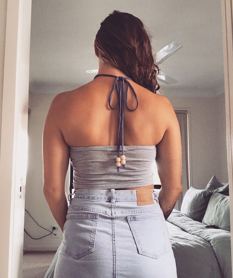May include: A woman wearing a gray crop top with a blue halter strap and wooden beads. The top is tied at the back of the neck. She is also wearing a light blue denim skirt.