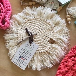 May include: Cream-colored, round macrame coaster with a fringed edge. The coaster is handmade and features a spiral pattern. A tag with the text "Gracy Mae Crochet" is attached. Other crochet items are visible in the background.