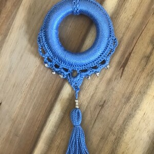 May include: A blue crocheted wreath with a tassel hanging from the bottom. The wreath is decorated with silver beads.