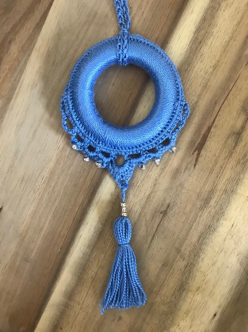 May include: A blue crocheted wreath with a tassel hanging from the bottom. The wreath is decorated with silver beads.