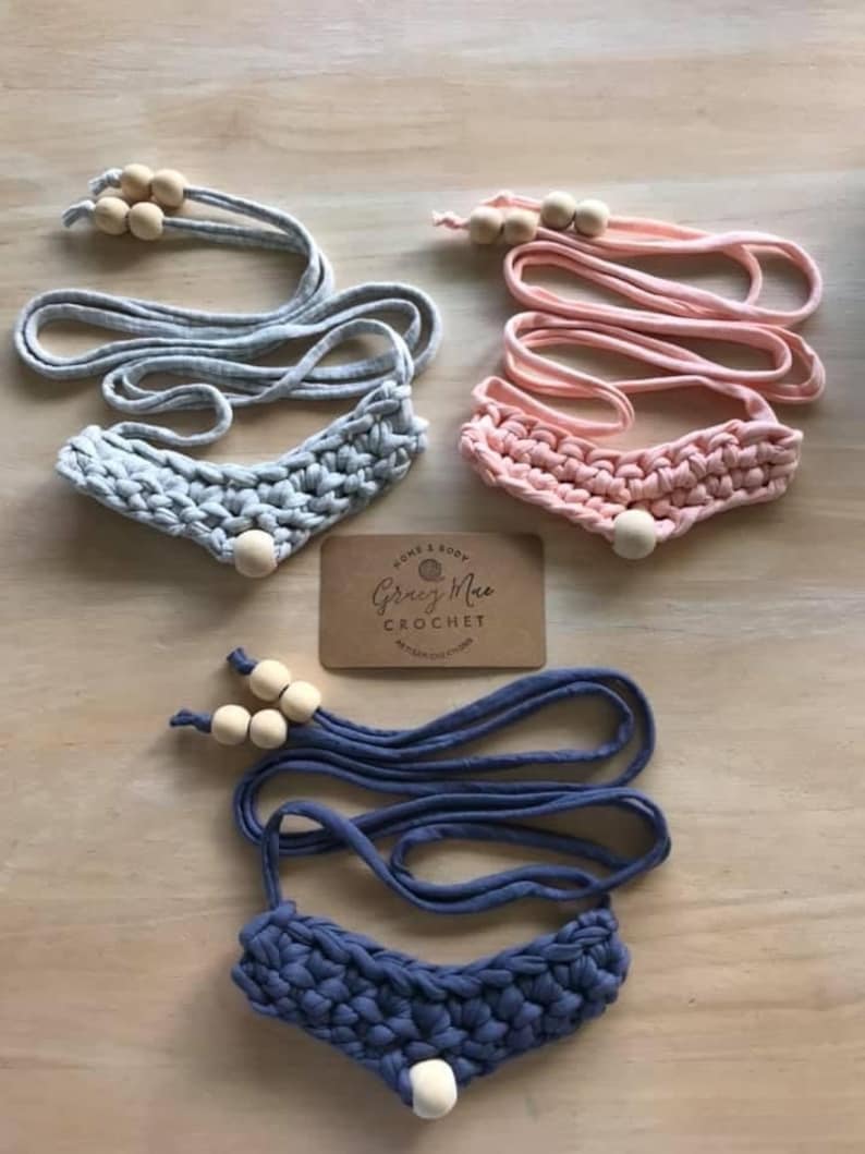 May include: Three crocheted necklaces with wooden beads. The necklaces are in gray, pink, and blue. The necklaces are handmade and have a simple design.