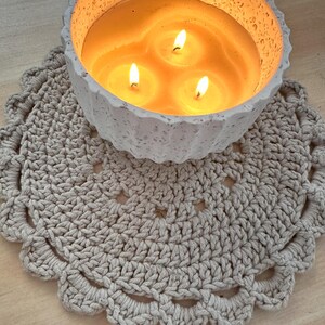 May include: A lit candle with three wicks in a speckled, light gray concrete bowl, resting on a beige crocheted doily. The candle's wax is a warm yellow, and the flames are bright.