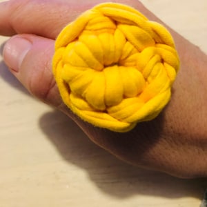 Puede incluir: Un anillo de crochet amarillo brillante hecho con restos de tela. El anillo se lleva en el dedo de una persona.