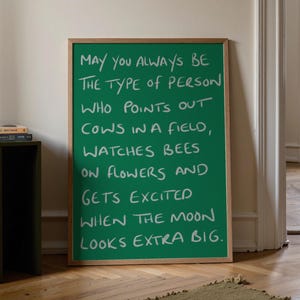 Puede incluir: Un cuadro rectangular con marco de madera clara que presenta un fondo verde sólido con texto blanco escrito a mano que dice: May you always be the type of person who points out cows in a field, watches bees on flowers and gets excited when the moon looks extra big.