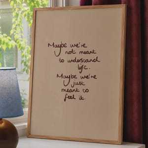 Op de afbeelding: Een rechthoekige ingelijste print met een lichtbeige achtergrond en donkerbruine handgeschreven tekst die luidt: Maybe we're not meant to understand life. Maybe we're just meant to feel it. De lijst is gemaakt van lichtgekleurd hout.