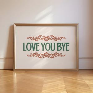Op de afbeelding: Een ingelijste print met de woorden "LOVE YOU BYE" in groen, met decoratieve bloemelementen in roodbruin. De lijst is goudkleurig en de print leunt tegen een witte muur op een houten vloer.