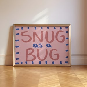 以下が含まれることがあります： 「SNUG as a BUG」という手書き風の文字が描かれた額入りのアートプリント。 ピンクと青の文字が、薄いピンク色の背景に映え、金色のフレームで縁取られています。 木の床の上に置かれています。
