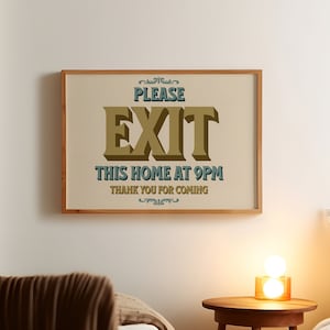 May include: A framed sign with the words "PLEASE EXIT THIS HOME AT 9PM THANK YOU FOR COMING" in a vintage style. The sign is in a light brown wooden frame and hangs on a white wall. A small wooden side table with a lamp is next to a bed.