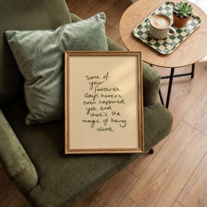 Minimalist typography print featuring the quote "Some of your favourite days haven't even happened yet and that's the magic of being alive" in an organic handwritten script. Warm beige background with olive green lettering creates a calming, hopeful and uplifting piece of inspirational wall art with a simple modern aesthetic.