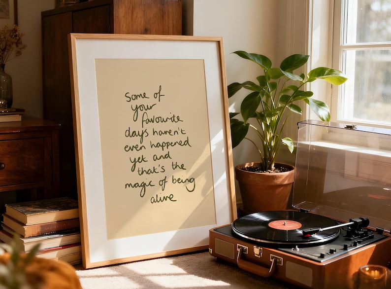 Minimalist typography print featuring the quote "Some of your favourite days haven't even happened yet and that's the magic of being alive" in an organic handwritten script. Warm beige background with olive green lettering creates a calming, hopeful and uplifting piece of inspirational wall art with a simple modern aesthetic.