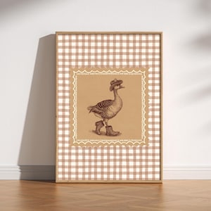 Op de afbeelding: Ingelijste kunstwerk met een sepia-getinte illustratie van een gans met een cowboyhoed en laarzen. Het kunstwerk staat tegen een bruine achtergrond met een decoratieve rand en een bruin-wit ruitpatroon.