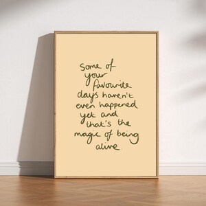 May include: A rectangular framed wall art piece featuring a light cream background with dark green handwritten text that reads: Some of your favourite days haven't even happened yet and that's the magic of being alive. The frame is light wood.