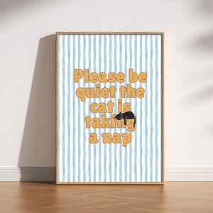 Könnte beinhalten: Gerahmter Druck mit hellblau-weiß gestreiftem Hintergrund. Der Text "Please be quiet the cat is taking a nap" steht in orangefarbenen Buchstaben. Eine schwarze Katzenillustration befindet sich in der Mitte des Textes.