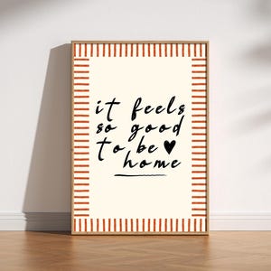Puede incluir: Una impresión artística enmarcada con el texto "it feels so good to be home" en escritura negra. La impresión tiene un fondo color crema con un borde a rayas rojo y crema y un marco de madera.
